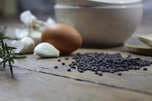 Close up detail of kitchen ingredients comprising of a brown egg, garlic, fresh rosemary and black lentils. All set on a landscape format against a wooden background.