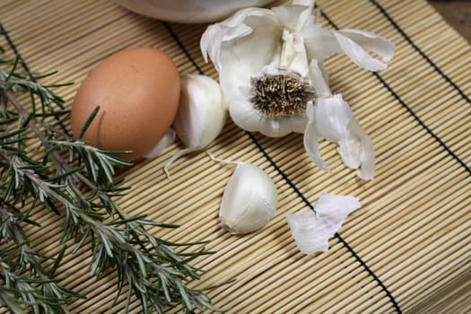 Close up detail of food ingredients comprising of a single brown egg, a bulb of garlic and a sprig of rosemary. All set on a landscape format on a bamboo matting base.