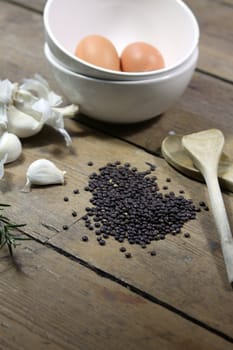 Kitchen ingredients comprising of brown eggs in a white bowl, garlic, fresh rosemary and black lentils. All set on a portrait format against a wooden background with two wooden spoons. Copy-space available.