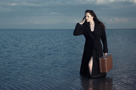 Woman in black coat standing in the sea with luggage