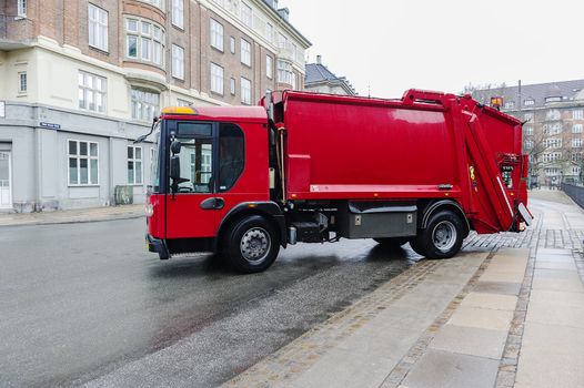 Red garbage disposal truck parked at the side of a street collecting household rubbish and waste for crushing, recycling and treatment or disposal on municipal dumps