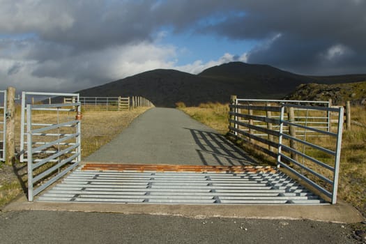 A metal cattle grid and gates with fences and a road leading towards the horizon with dark mountains.