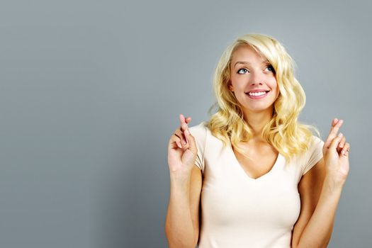 Smiling blonde caucasian woman with fingers crossed on gray background