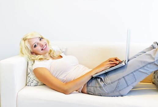 Smiling young blonde woman using laptop computer laying on couch
