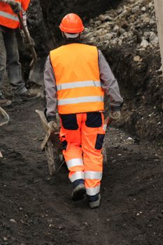 Worker wearing orange uniform pushing wheelbarrow to another worker on the construction site