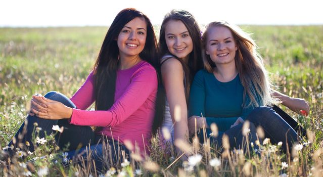 three young beautiful woman sitting in a field on the  sky background 