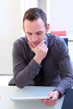 Young professional business man concentrating and thinking with his chin resting on his hand as he reads the screen of his tablet computer
