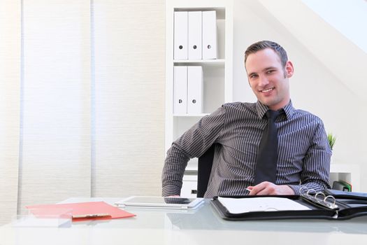 Smiling successful young businessman sitting at his desk which is spread with folders and a tablet computer