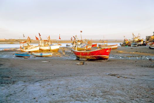 Dwarka Roadtrip. Landscape of an orange evening sun falling on banked and grounded fishing boats and ferries in Gujarat India. Hazy blue sky and silty foreground with motorbike tire marks
