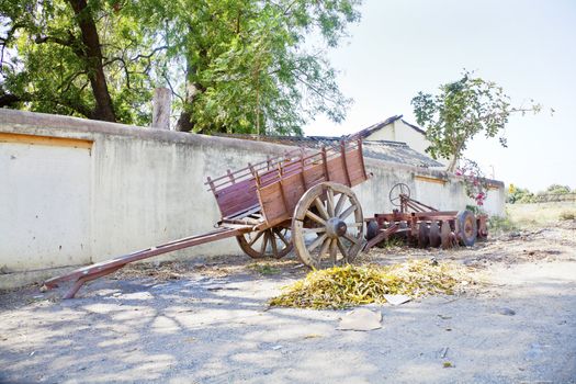 Landscape at the outskirts of an Indian hinterlands village of a wall lined with farm machinery and parked bullock cart under the shade of trees