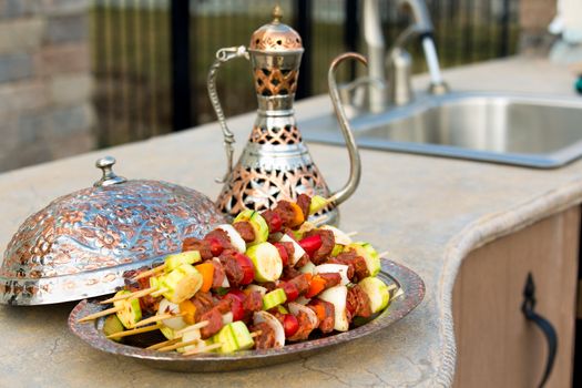 Raw meat and vegetable skewers are waiting on the outdoor kitchen counter top. Skewers are in the copper plate  along with complimenting ornamented copper pitcher.