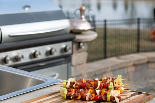 Seasoned meat skewers with vegetables on multicolored cutting board, on outside kitchen counter top next to the barbecue.