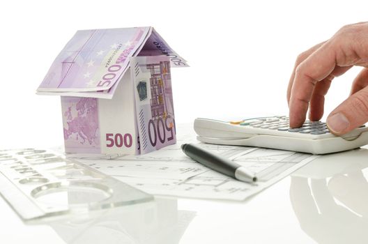 Architect calculating costs of house construction with blueprints and house made of money on his desk. Over white background.