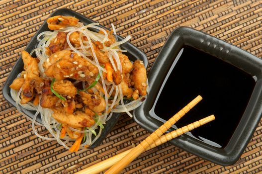 Chicken Teriyaki with Chinese Glass Noodles and Soy Sauce in Black Square Shape Bowls with Pair of Chopsticks on Straw mat background. Top View