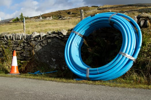 A coiled blue water pipe leaning on a wall on the side of a road with a red and white marker cone.