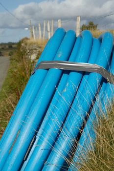 A coiled blue water pipe leaning on a wall on the side of a road verge with a fence and blue cloudy sky.