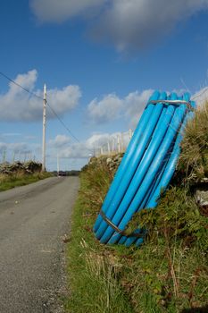 A coiled blue water pipe leaning on a wall on the side of a road verge with a fence and blue cloudy sky.