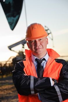 Oil worker in orange uniform and helmet on of background the pump jack and sunset sky.