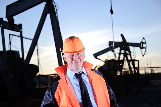 Oil worker in orange uniform and helmet on of background the pump jack and sunset sky.