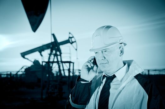 Oil worker in uniform and helmet, with mobile phone on of background the pump jack and sky. Toned.