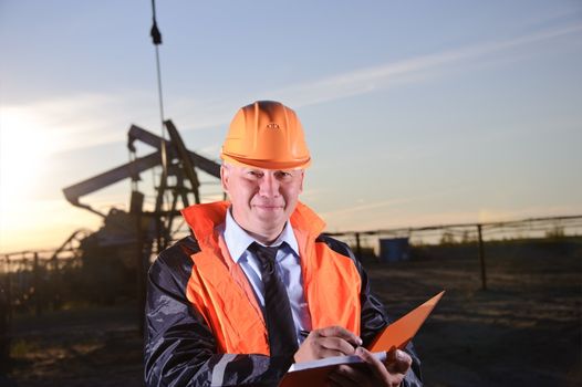 Oil worker in orange uniform and helmet on of background the pump jack and sunset sky. Look into the camera.