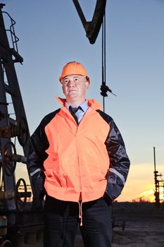 Worker in orange uniform and helmet on of background the pump jack and sunset sky. Severe. Hands in pockets.