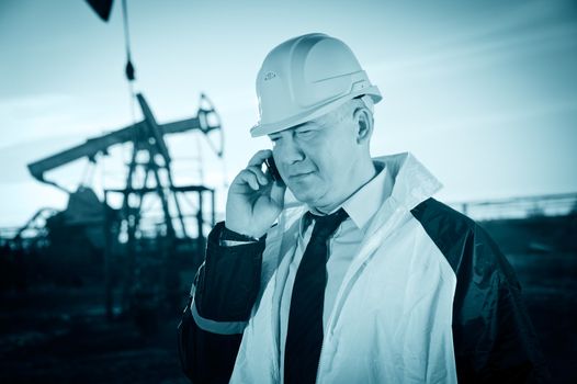 Oil worker in uniform and helmet, with mobile phone on of background the pump jack and sky. Toned.