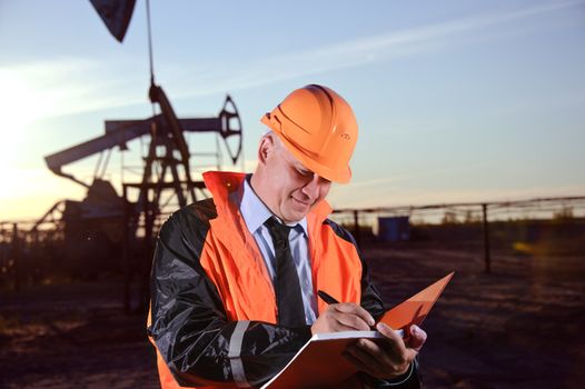 Oil worker in orange uniform and helmet on of background the pump jack and sunset sky.