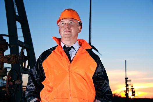 Oil worker in orange uniform and helmet on of background the valves, piping and sunset sky.