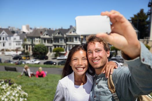 Happy young couple in San Francisco Alamo Square taking self-portrait photo pictures with smart cell phone camera. Multiethnic interracial couple tourists on sightseeing travel in California, USA