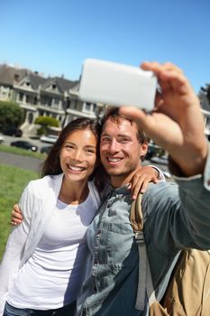 Couple fun taking self-portrait photo with cell phone camera. Multiracial happy tourist couple on travel vacation in San Francisco, Alamo Square, USA.