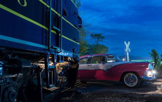 Blue train diesel railway engine almost hits vintage thunderbird car at railroad crossing at night