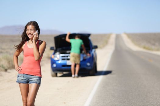 Car breakdown - woman phone calling auto service. Positive happy smiling woman on smart mobile phone, with man looking at car engine in background on highway in California, USA. Young couple on road.
