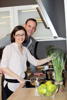 Young couple preparing dinner standing close together over the stove looking at the camera with cheerful smiles