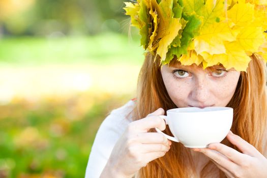 young redhead teenager woman in a wreath of maple leaves with cup of tea