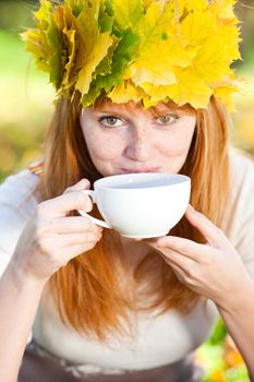 young redhead teenager woman in a wreath of maple leaves with cup of tea