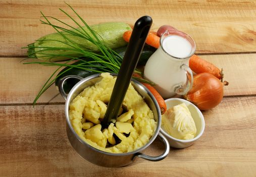 Preparing Vegetables Puree. Potato Masher in Potato, Jar of Milk, Butter, Onion, Carrot, Chive and Zucchini closeup on Wooden background
