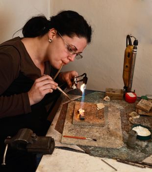 Female jeweller blowing a torch to melt metal for making a brooch in her workshop.