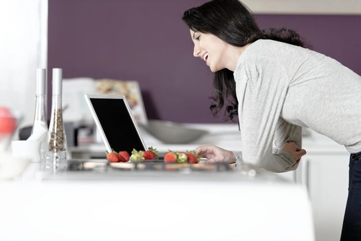 Beautiful young woman reading a recipe from a laptop in her kitchen