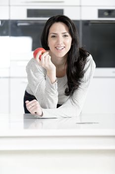 Beautiful young woman eating an apple in her white kitchen relaxing