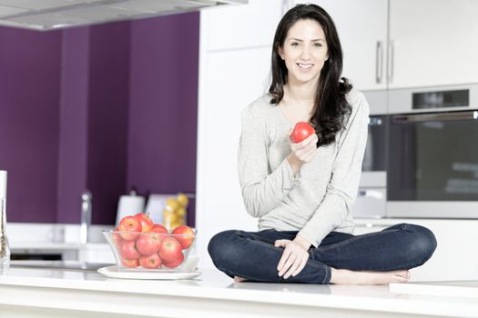 Beautiful young woman eating an apple in her white kitchen relaxing