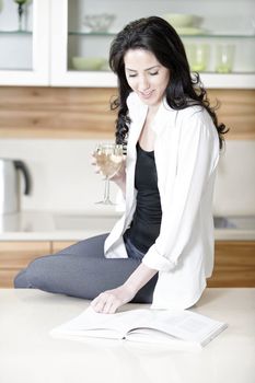 Attractive young woman reading a recipe book while enjoying a glass of wine in the kitchen