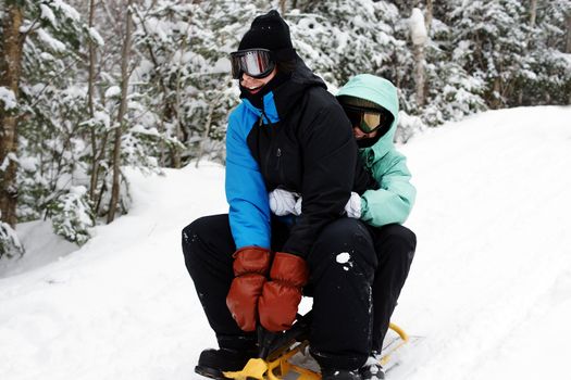 Young man and woman having fun and laughing, in full winter apparel including ski mask, sliding down a hill on a small three skis, great for nature, outdoors or seasonal concept.