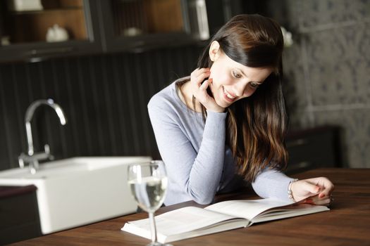 Attractive young woman reading a recipe from a cookery book in her kitchen with a glass of wine.