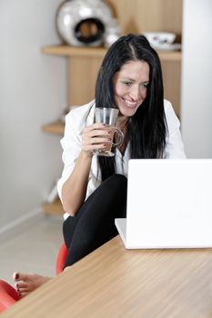 Attractive young woman taking a coffee break in her kitchen with her laptop.