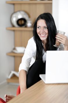 Attractive young woman taking a coffee break in her kitchen with her laptop.