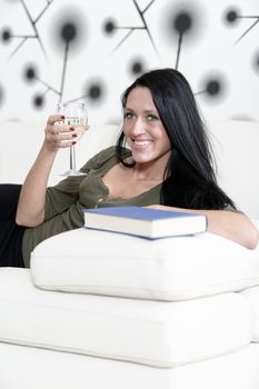 Attractive young woman lying on her sofa enjoying a glass of wine and a good book.