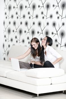 Two attractive young friends catching up at home on the sofa with a glass of wine using a laptop computer.