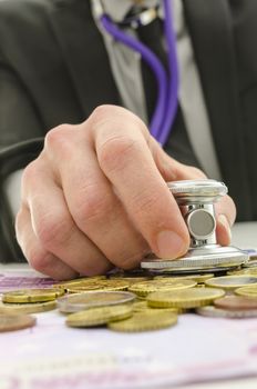 Male hand holding stethoscope over Euro coins and banknotes.