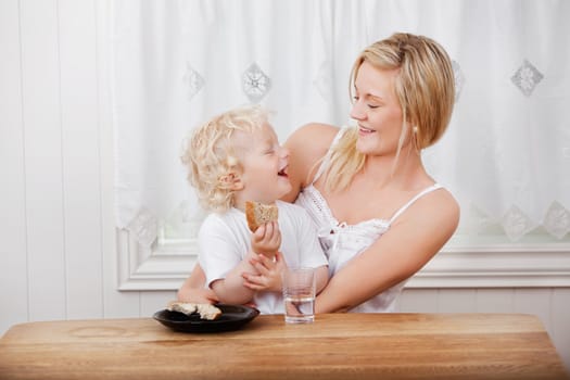 Young beautiful mother looking at cheerful baby boy eating bread at dining table